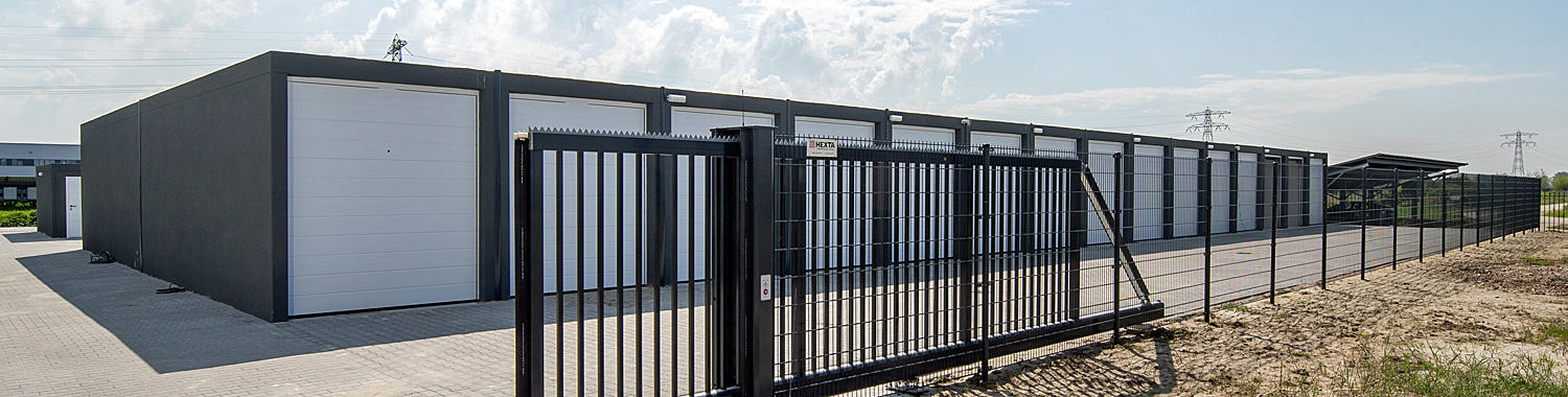 Eine reihe von dunkelgrauen Fertiggargen mit weißen Toren die teil eines Garagenhofes sind. Davor ist ein Zaun mit einem großen elektrischen Tor , welches der Zugang zu dem Gelände des Garagenparks ist. 